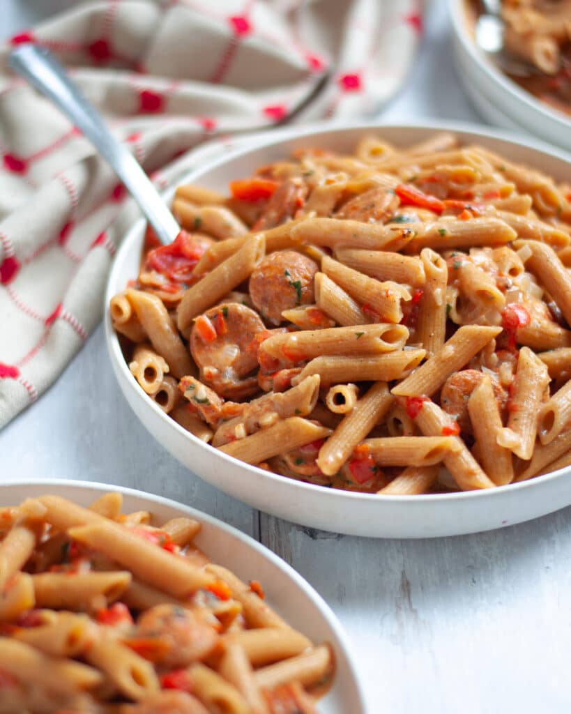 close up of a serving bowl filled with this chicken andouille sausage cajun pasta. plates filled with pasta surround the serving dish as well as a red and tan linen.