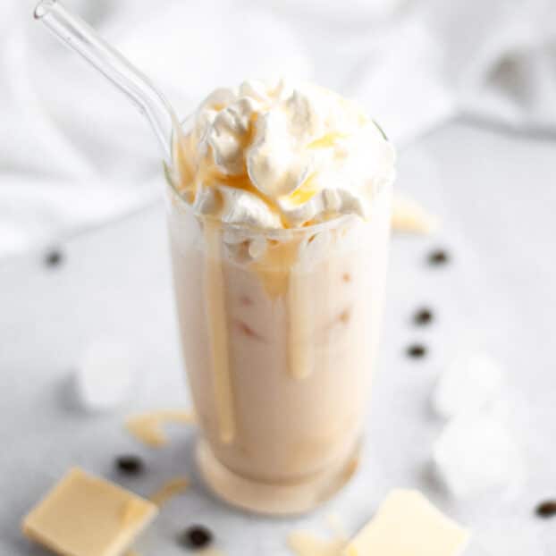 close up of a tall glass filled with a white iced mocha that is topped with a generous amount of whipped cream and white chocolate syrup that is dripping down the glass. The drink is surrounded by white chocolate squares, coffee beans, ice, and a white linen.
