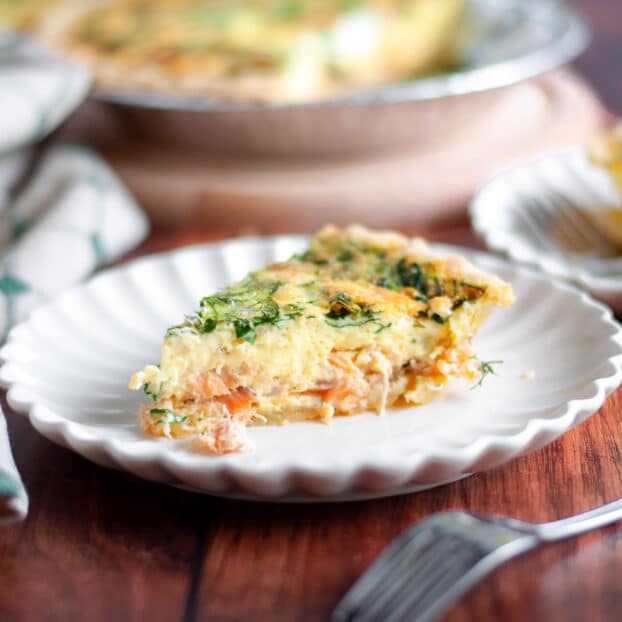 a slice of smoked salmon and spinach quiche on a serving plate is the focus on this image. Additional serving plates and the pie pan of spinach salmon quiche are in the background, along with a pie server and tan and green linen.