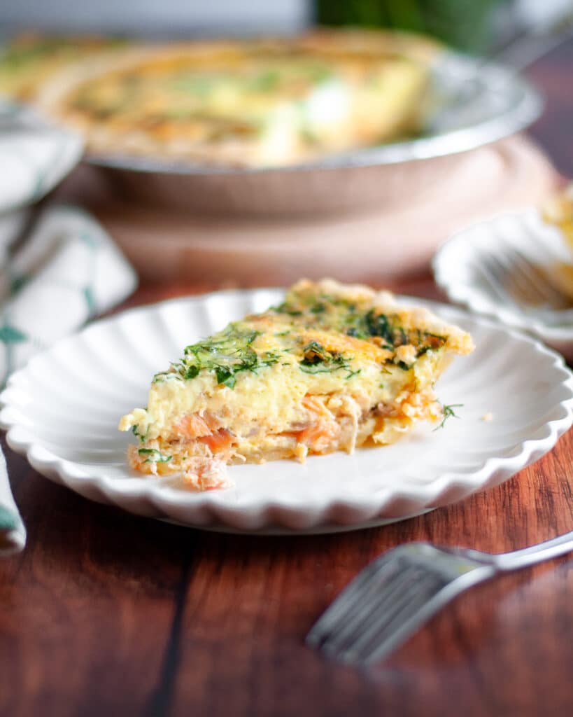 a slice of smoked salmon and spinach quiche on a serving plate is the focus on this image. Additional serving plates and the pie pan of spinach salmon quiche are in the background, along with a pie server and tan and green linen.