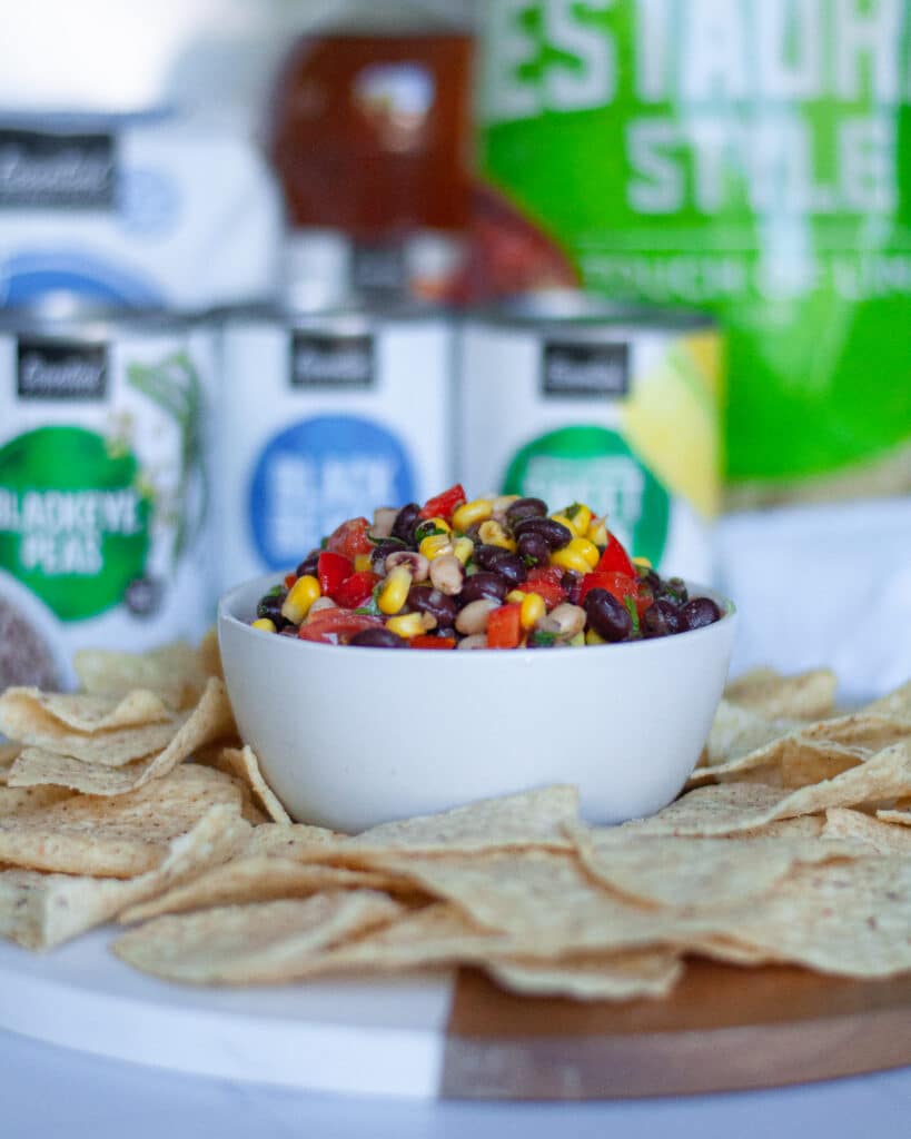 bowl of texas caviar on a board surrounded by tortilla chips. In the background, there are packages of ingredients.