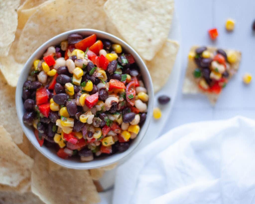 top down view of a bowl of cowboy caviar on a board surrounded by tortilla chips. There is a chip full of dip and a white linen as well.