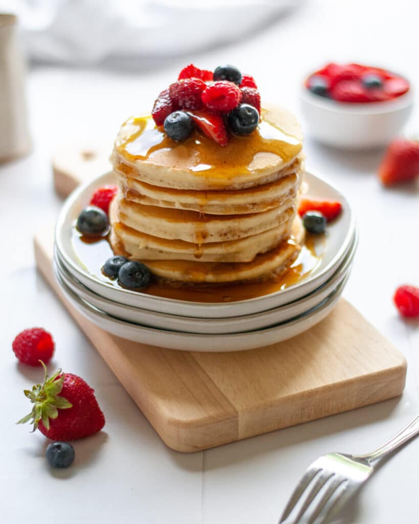 hero shot of a stack of oatmilk pancakes covered in berries. The plate is surrounded additional plates of pancakes, berries, and a carafe of maple syrup.