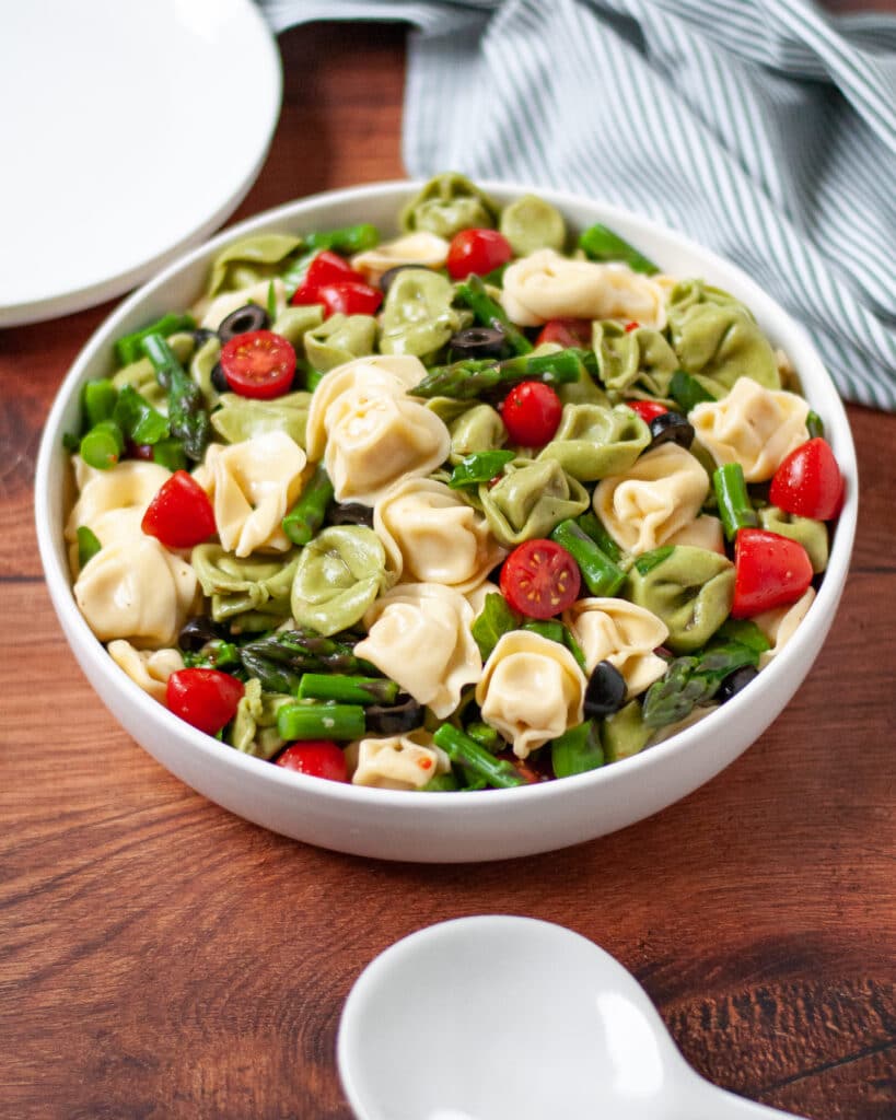 close up of a serving bowl filled with summer tortellini salad. a striped linen and serving plates peak into the side and back of the photo, with a serving spoon sitting in front of the bowl.