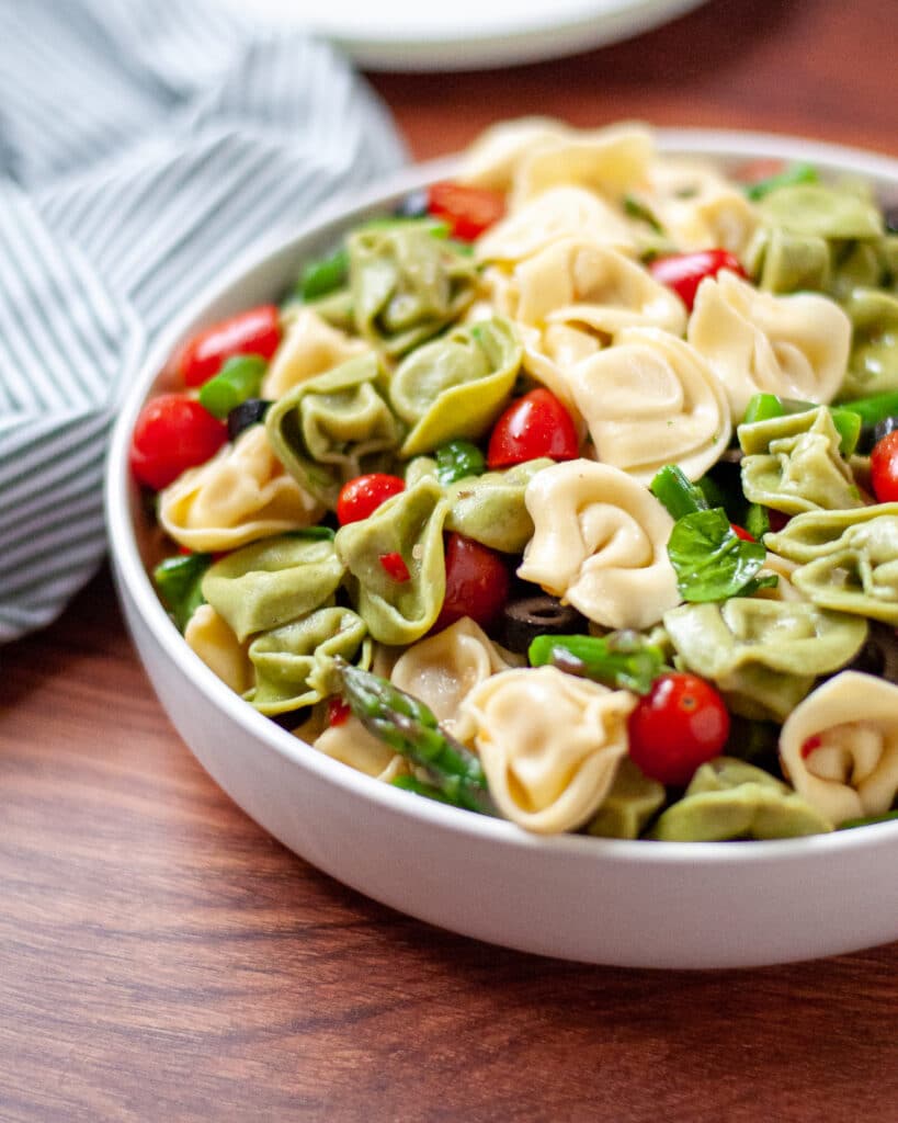 close up of a serving bowl filled with summer tortellini salad. a striped linen and serving plates peak into the side and back of the photo.