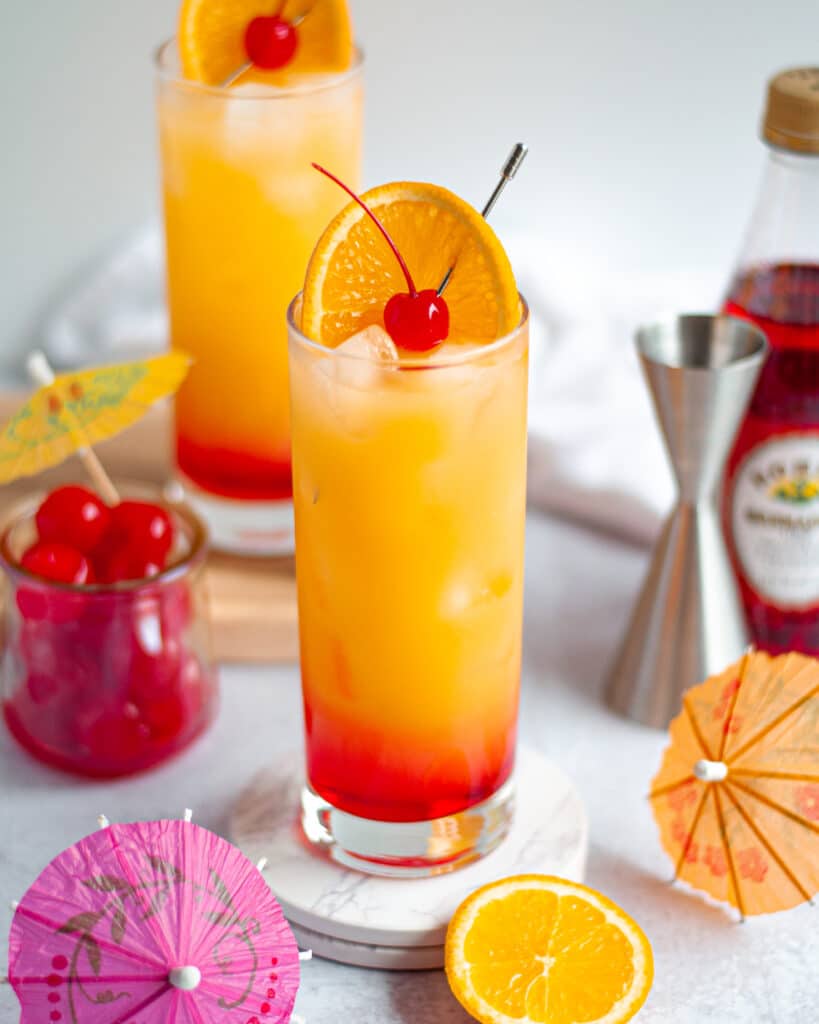 Close up of a highball filled with this vodka sunrise recipe and garnished with an orange slice and maraschino cherry. The glass is surrounded by drink umbrellas, more cherries, orange slices, a jigger, grendadine, with another vodka sunrise cocktail in the background.