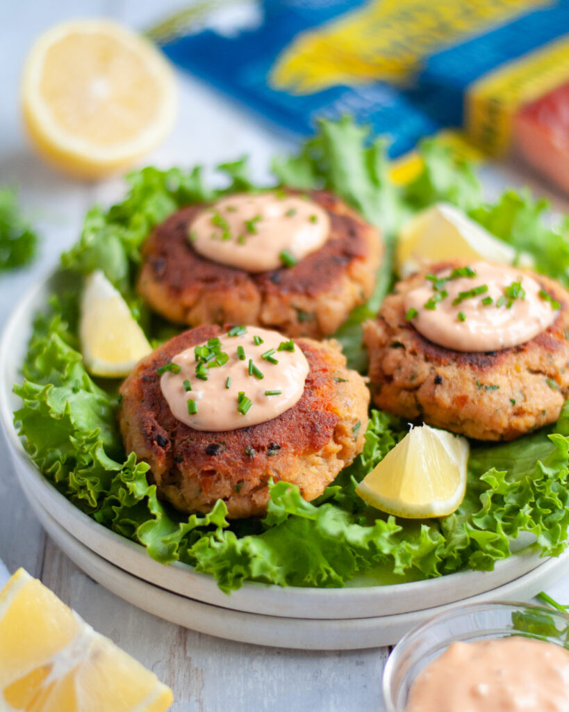 close up of a plate of smoked salmon patties, topped with a sauce and fresh chives. The smoked salmon cakes are sitting on leaves of lettuce with lemon wedges.