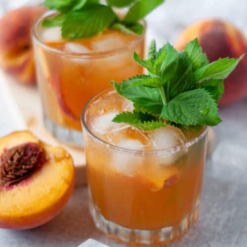 close up of a low ball glass filled with this peach whiskey smash recipe. The glass is garnished with a large sprig of fresh mint. The glass is surrounded by another bourbon smash cocktail, fresh peaches, and ice cubes.