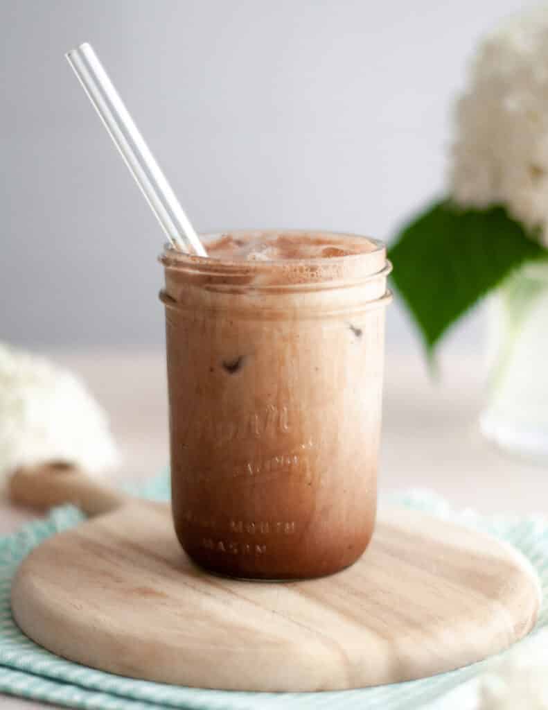close up of an iced chocolate almondmilk shaken espresso in a jar with a glass straw. The shaken espresso is sitting on a wooden board on top of a blue and white checkered napkin which is surrounded by hydrangea flowers.