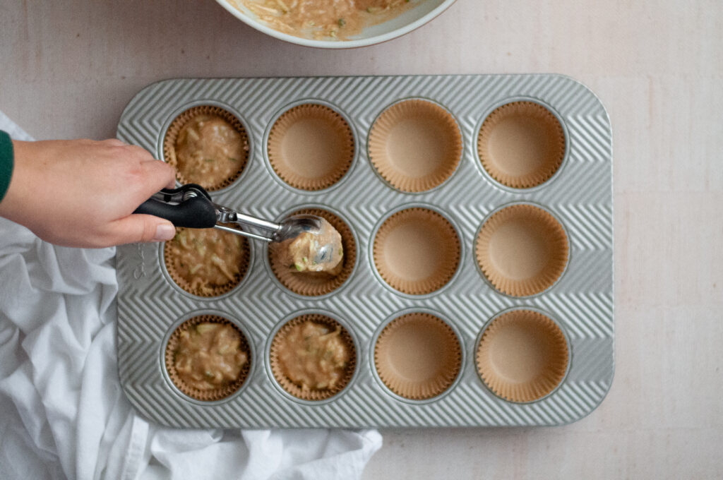 process shot showing a large cookie scoop being used to fill a muffin tin filled with liners.