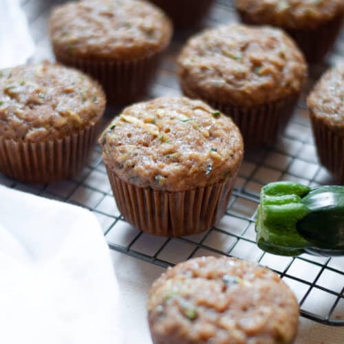 close up of a zucchini applesauce muffin sitting on a wire cooling rack, along with the additional muffins from the batch., a white linen, and zucchinis