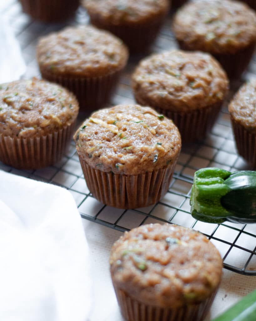 close up of a zucchini applesauce muffin sitting on a wire cooling rack, along with the additional muffins from the batch., a white linen, and zucchinis
