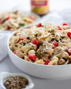 close up of a serving dish filled high with pesto pasta salad with chicken. The bowl is surrounded by a white linen, plate of pasta, small bowl of pesto, and jar of pesto.