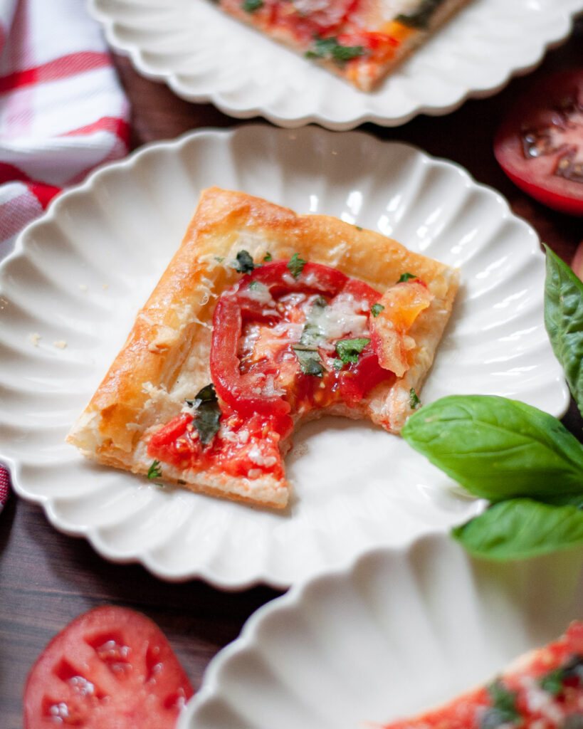 close up of a slice of tomato tart on a white plate. the tart is surrounded by other plates with tarts, a red and white checkered linen, slices of tomatoes, and a sprig of fresh basil.