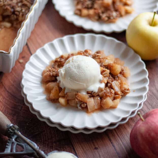 close up of a serving of apple crisp on a delicate white plate topped with a scoop of vanilla ice cream. Another serving of apple crisp with oat topping is shown in the background as well as the 9x13 pan filled with the rest of the crisp.