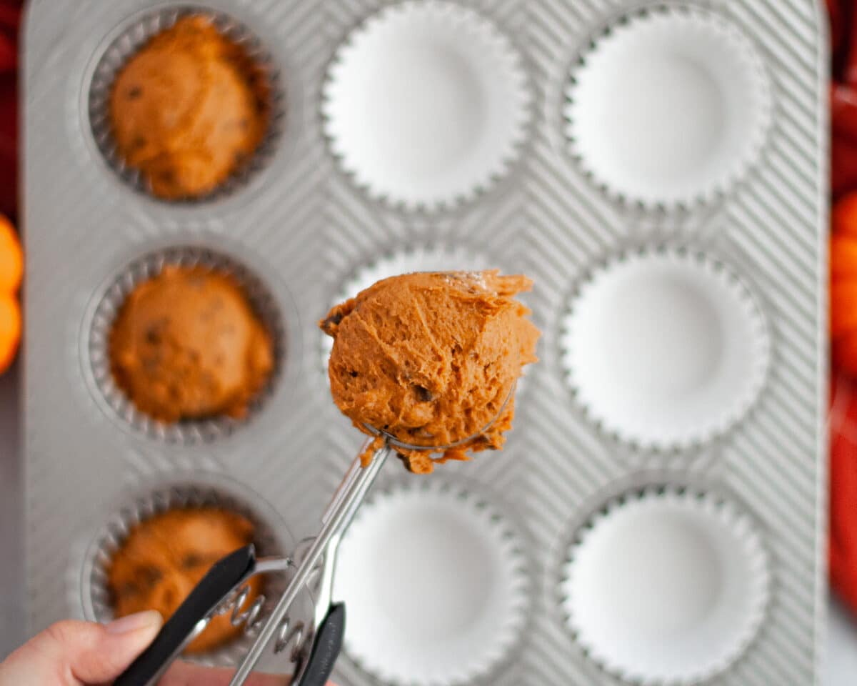 a large cookie scoop filled with pumpkin muffin batter above a silver muffin tin with white liners. 3 of the tins have already had batter scooped into them.