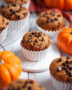 a pumpkin chocolate chip muffin in a white liner sitting on a white coaster. The muffin is surrounded by additional 3 ingredient pumpkin muffins, mini pumpkins, and an orange linen.