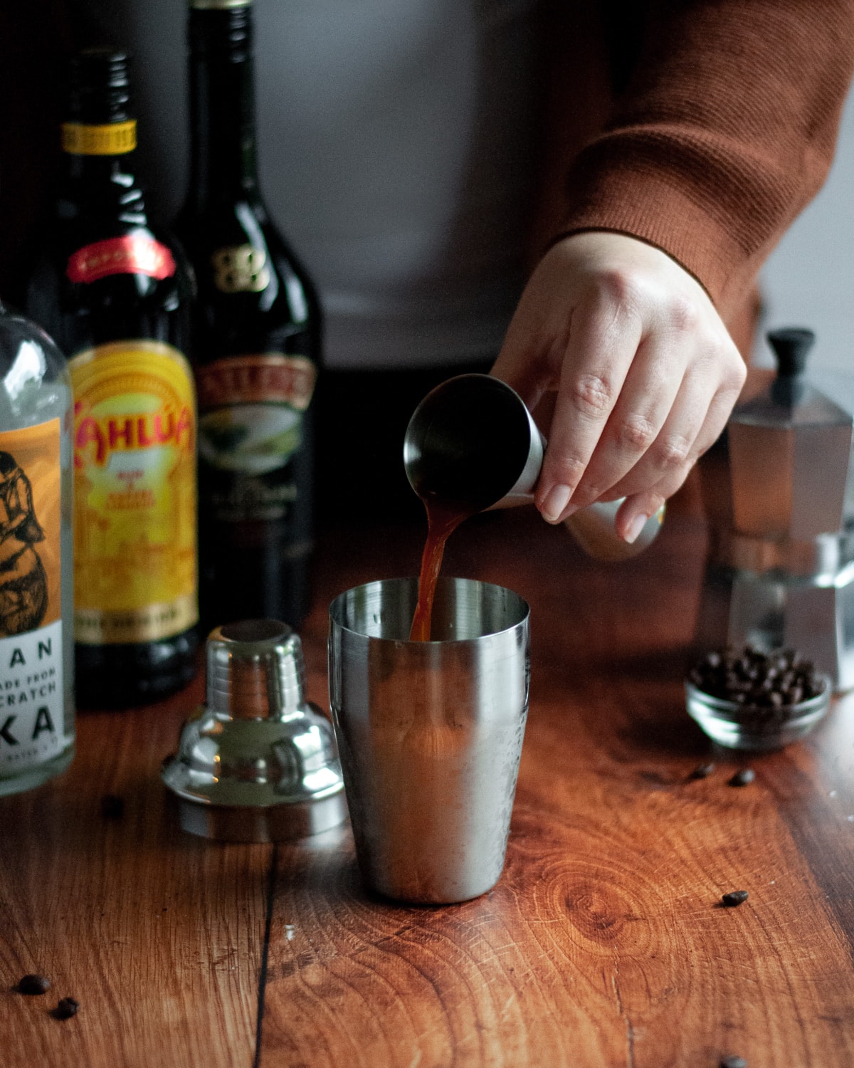 process shot of making an espresso martini with baileys. a person is pouring one of the liquid ingredients into a silver cocktail shaker from a jigger. liquor bottles, a moka pot, and coffee beans sit in the background.