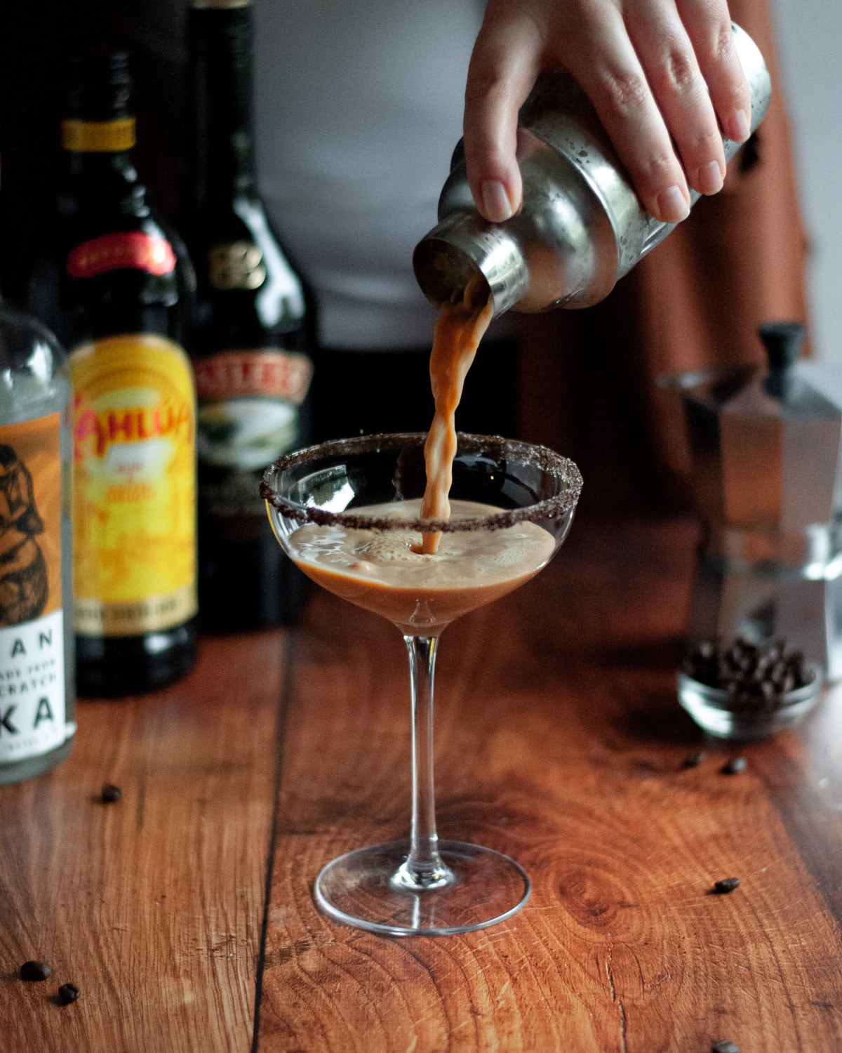 process shot of making an espresso martini with baileys. a person is pouring a freshly shaken cocktail into a coupe glass with a coffee-sugar rim. liquor bottles, a moka pot, and coffee beans sit in the background.