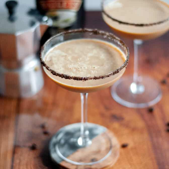 a coupe glass with a coffee and sugar rim filled with a baileys espresso martini. a bowl of coffee beans is in the foreground with additional coffee beans scattered around the scene. another cocktail, a silver moka pot, and a bottle of baileys irish cream are in the background.