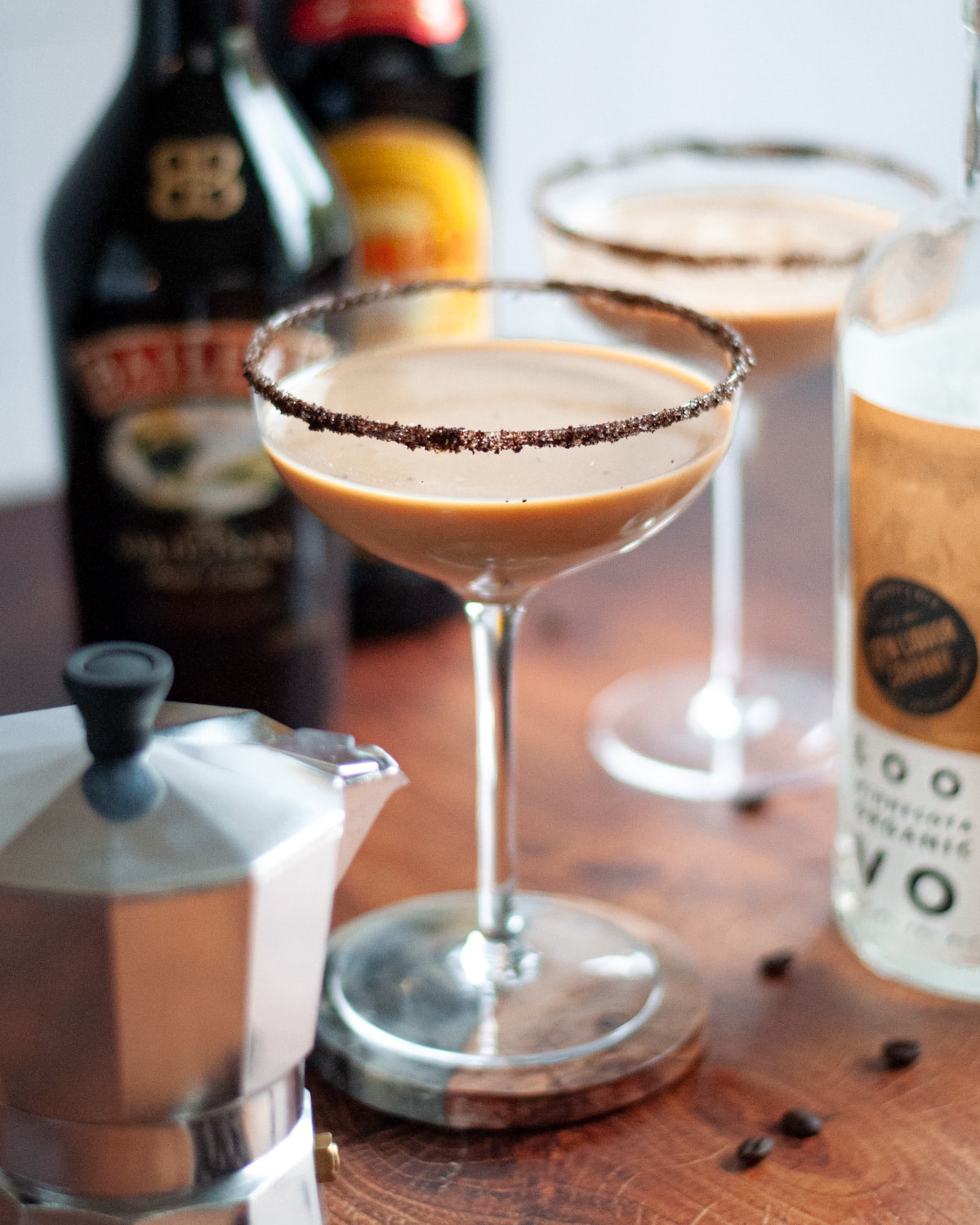 a coupe glass with a coffee and sugar rim filled with a baileys espresso martini. a bowl of coffee beans is in the foreground with additional coffee beans scattered around the scene. another cocktail, liquor bottles, and a moka pot surround the cocktail.