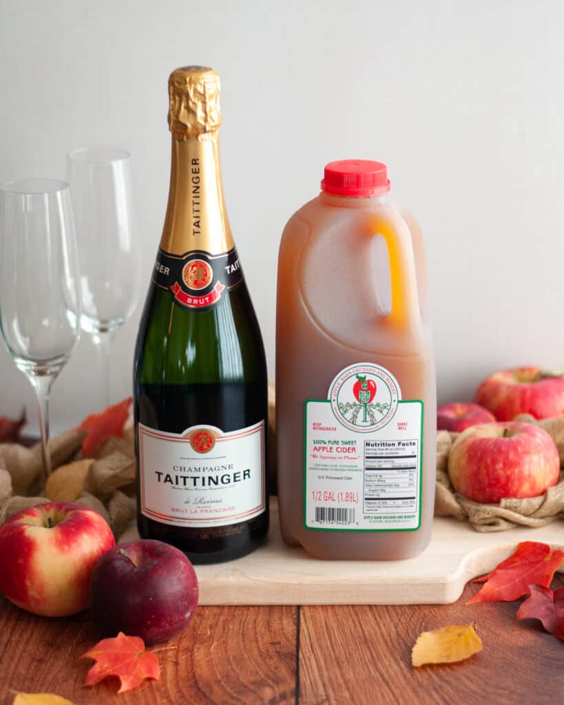 ingredients needed to make an apple cider mimosa. the bottle of champagne and container of apple cider is surrounded by apples, fall leaves, a gold linen, and champagne flutes.