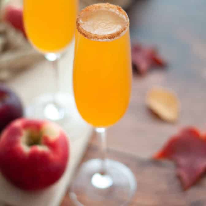 close up of an apple cider mimosa with a cinnamon-sugar rim. The champagne flute is surrounded by apples, fall leaves, and a golden linen.