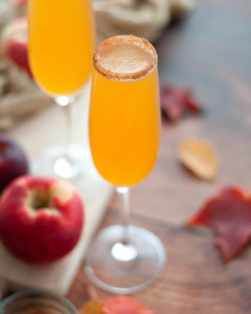 close up of an apple cider mimosa with a cinnamon-sugar rim. The champagne flute is surrounded by apples, fall leaves, and a golden linen.