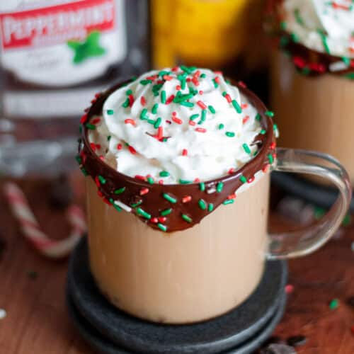 a mug of boozy christmas coffee sitting in front of liqueur bottles. the rimmed with chocolate and sprinkles, and is surrounded by coffee beans, sprinkles, chocolate pieces, and candy canes surround the mug.