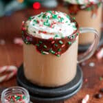 a clear mug rimmed with chocolate and sprinkles filled with boozy christmas coffee. the coffee is topped with whipped cream and sprinkles. coffee beans, sprinkles, chocolate pieces, and candy canes surround the mug.