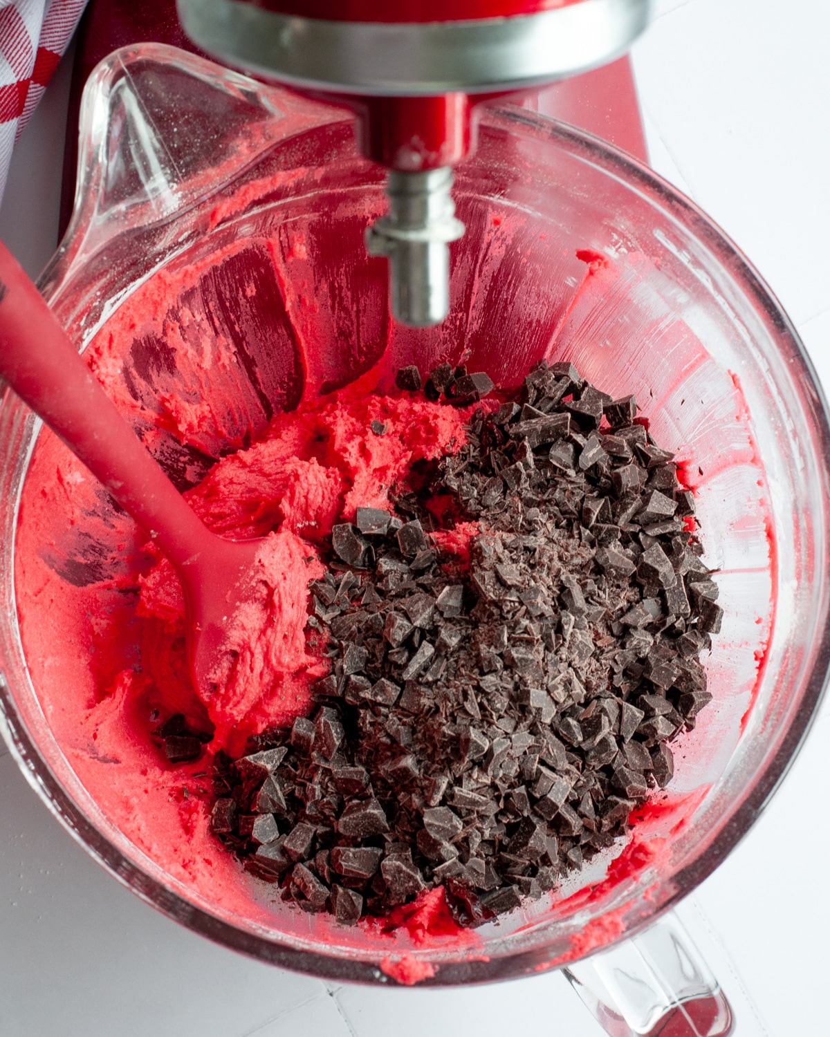 a glass mixing bowl filled with bright red dough and chopped pieces of chocolate ready to be mixed in. the mixing bowl sits in the base of a red stand mixer and a red and white checkered linen is in the background.