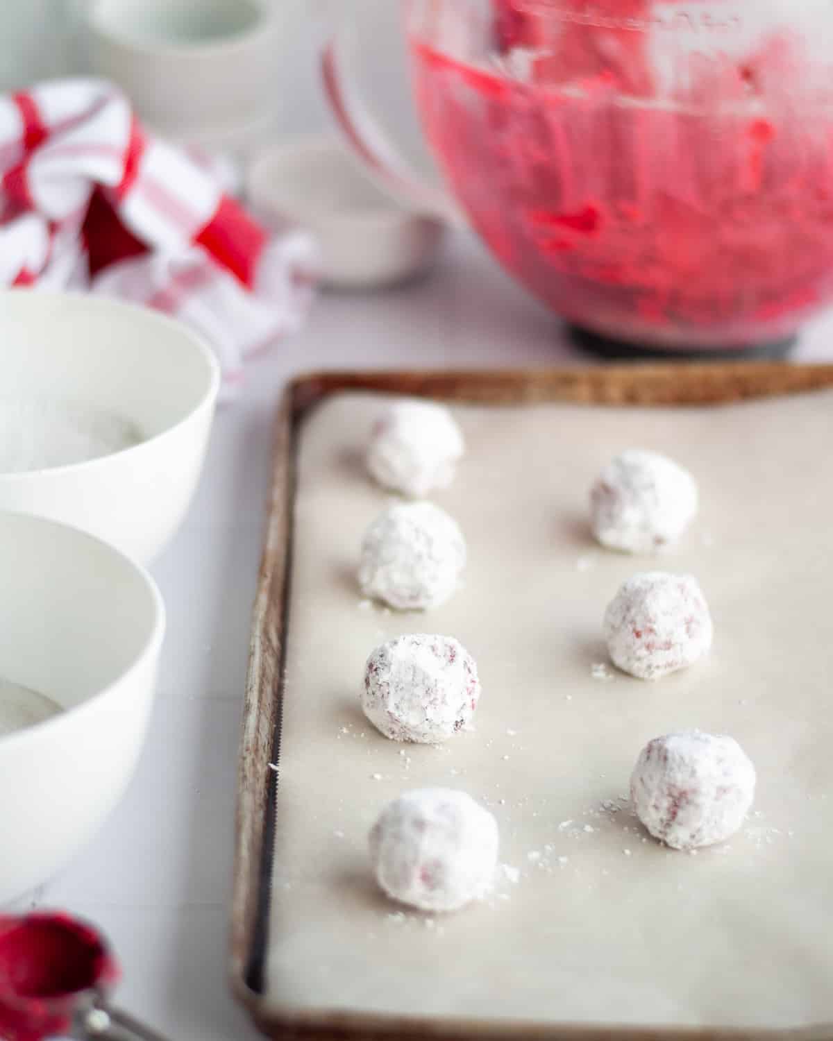 baking sheet lined with parchment paper and filled with freshly rolled chocolate cherry crinkle cookie dough balls. bowls of granulated sugar and powdered sugar peak into the picture showing what the dough balls are rolled in. the mixing bowl of bright red dough sits in the background.