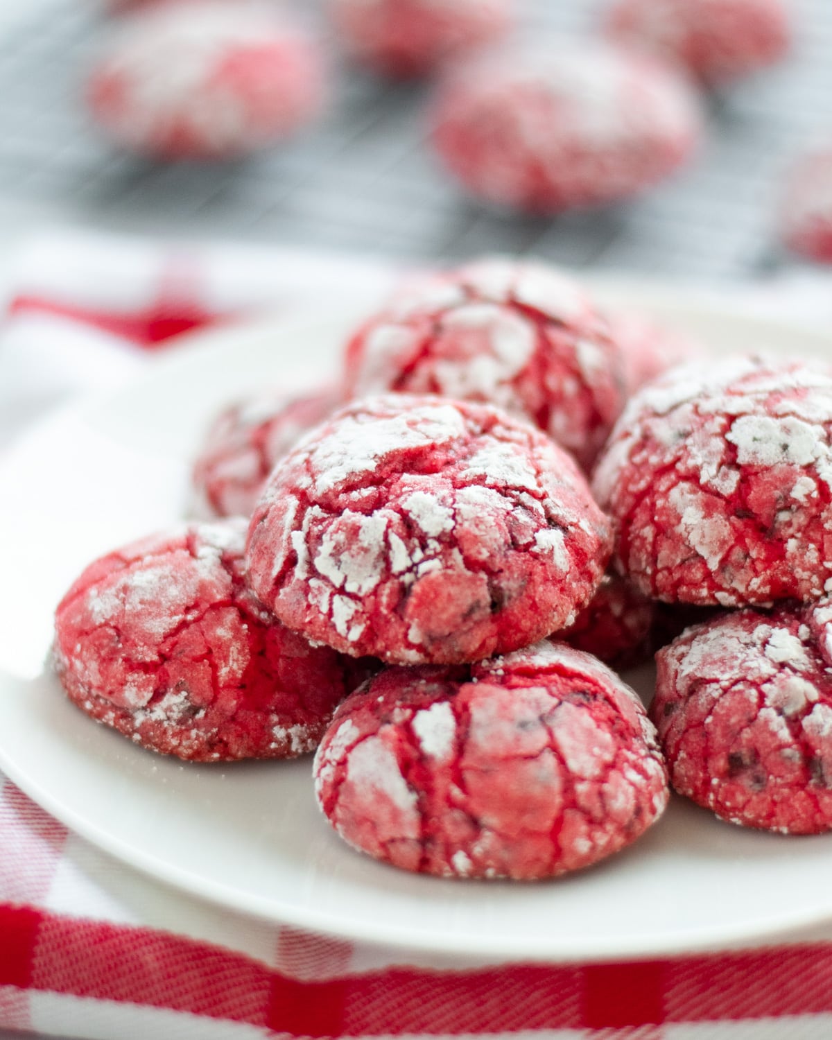 white plate full of red and white chocolate cherry crinkle cookies set on a red and white checkered linen. Blurred in the background you can see a wire cooling rack with additional cookies.