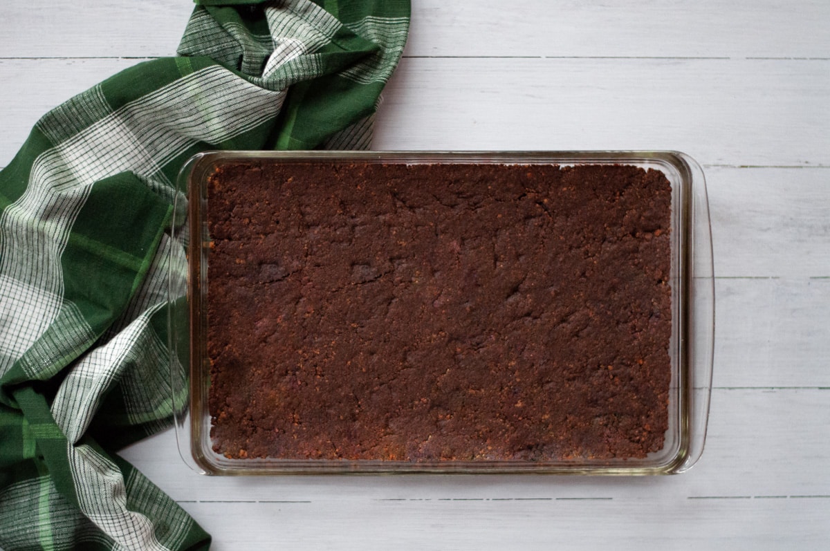The base layer of chocolate mint bars pressed into a glass 9x13 pan. The pan sits next to a green and white plaid linen.