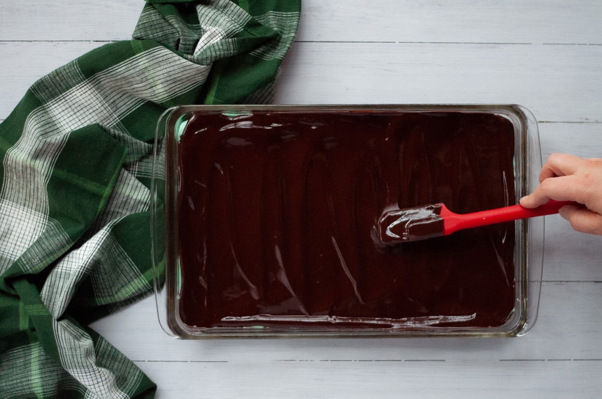 A person spreading the chocolate ganache top layer over the set mint middle layer in a 9x13 pan. The pan sits next to a green and white plaid linen.