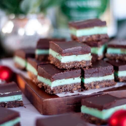 Cut chocolate mint bars sitting in a layer on a dark wooden board. Christmas ornaments, a green plant in a silver container, and bottle of creme de menthe surround the bars.