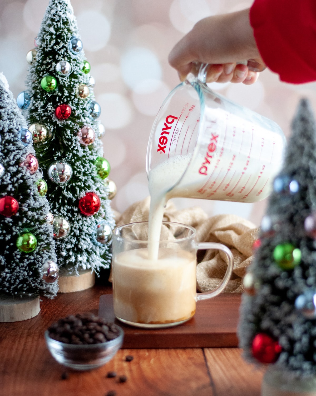 process shot for making an eggnog latte. shows frothed eggnog and milk being poured from a liquid measuring cup into a clear glass mug. the mug is on a wooden board and surrounded by miniature christmas trees with multi-colored ornaments, a golden linen, a small bowl of coffee beans, and a golden, twinkling background.