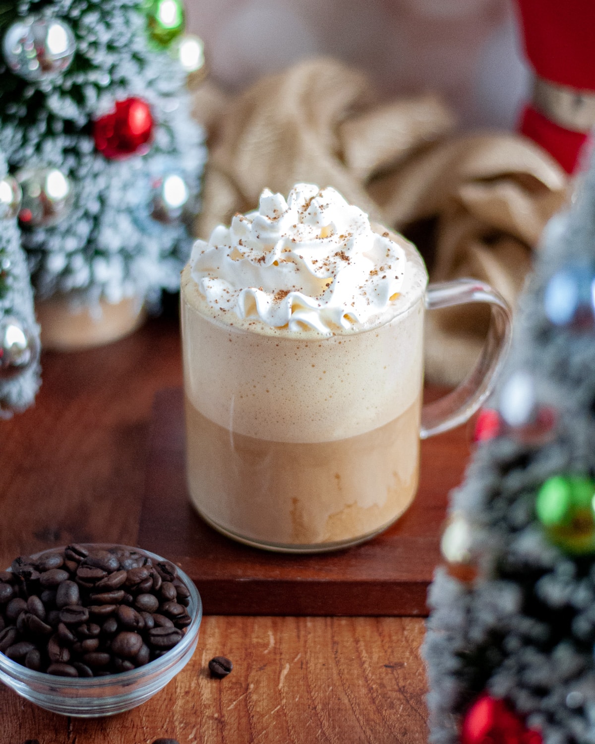 an eggnog latte sitting on a wooden board. the mug is surrounded by miniature christmas trees with multi-colored ornaments, a golden linen, a small bowl of coffee beans, a red moka pot, and a golden, twinkling background.