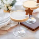 an eggnog cocktail with rum in a textured coupe glass with a gingerbread star cookie for garnish. another cocktail sits in the background on a wooden board with a white holiday plant and white linen.