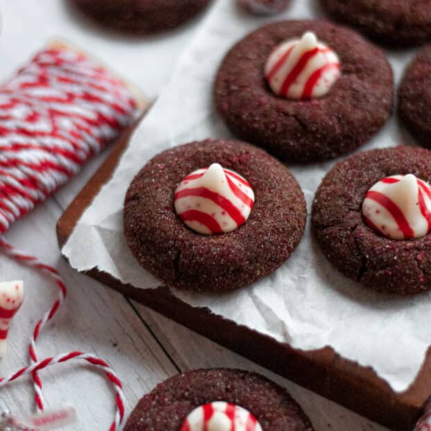 Chocolate peppermint kiss cookies sitting on a sheet of parchment paper on a dark wooden board. A spool of red and white twine sits nearby, as well as more cookies and candy cane kisses.