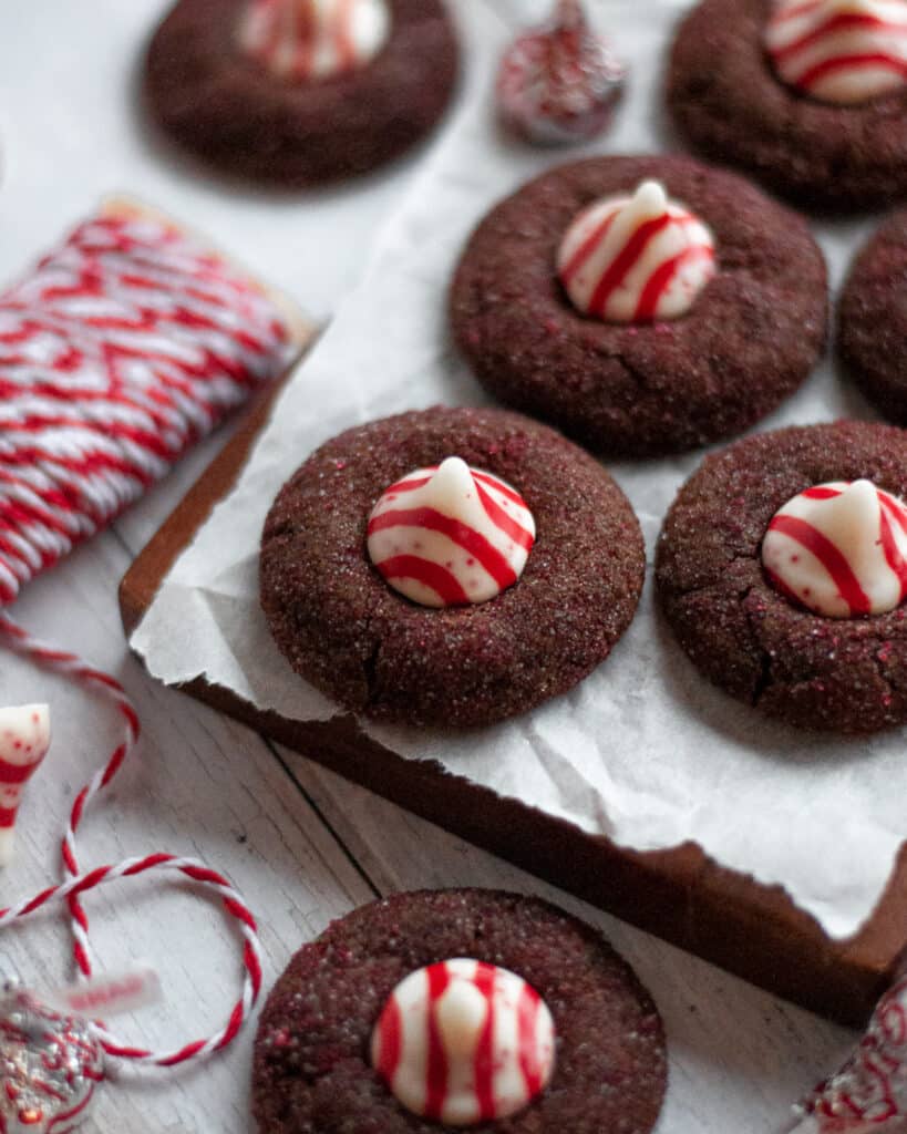 Chocolate peppermint kiss cookies sitting on a sheet of parchment paper on a dark wooden board. A spool of red and white twine sits nearby, as well as more cookies and candy cane kisses.