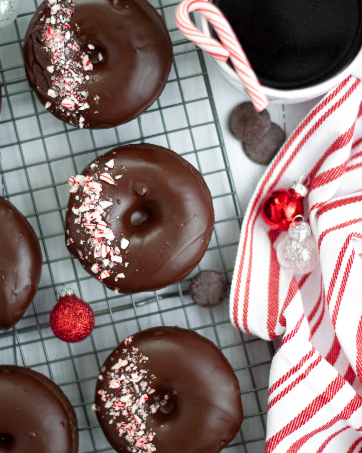 peppermint mocha donuts sitting on a wire cooling rack, cooled and ready to eat. the donuts are surrounded by red and white christmas ornaments, pieces of chocolate, a red and white striped linen, candy canes, and a mug of coffee.