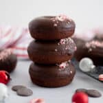 a stack of 3 peppermint mocha donuts surrounded by candy canes, chocolates, christmas ornaments, and a red and white linen.