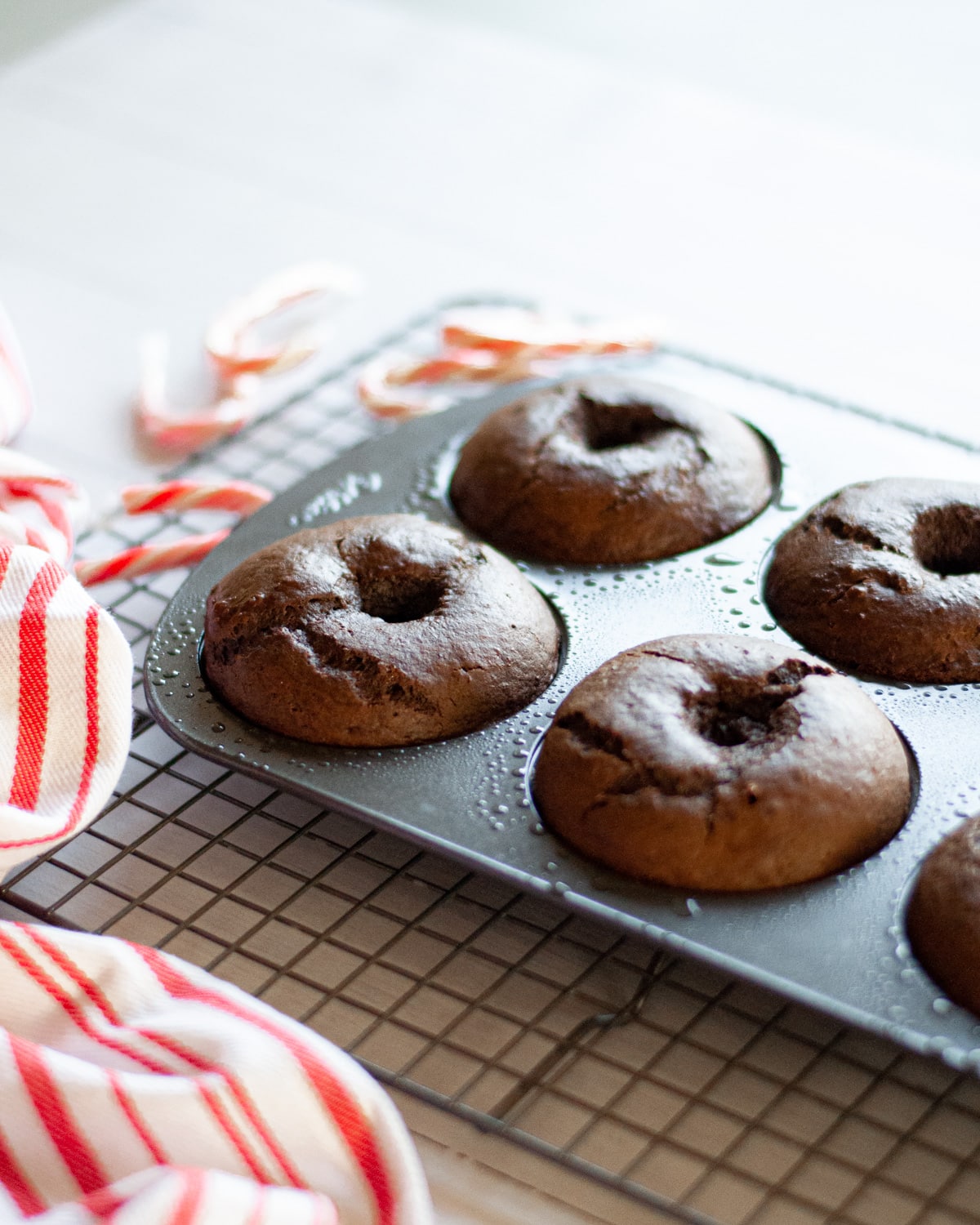 freshly baked chocolate donuts in a donut pan on a wire cooling rack. candy canes and a red and white linen sit near the pan.