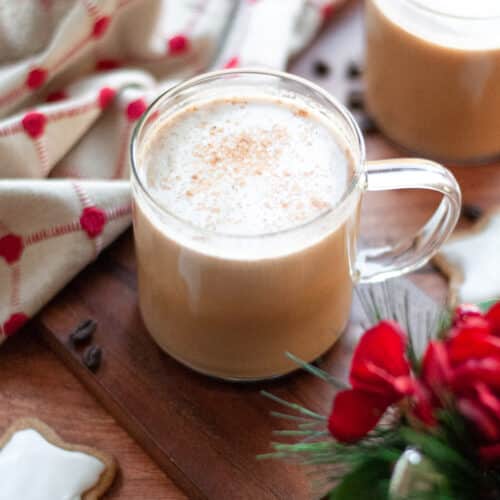 a glass mug with a homemade gingerbread latte sitting on a wooden board. Another mug sits in the background, along with a linen, coffee beans, an iced gingerbread cookie, and a holiday plant.