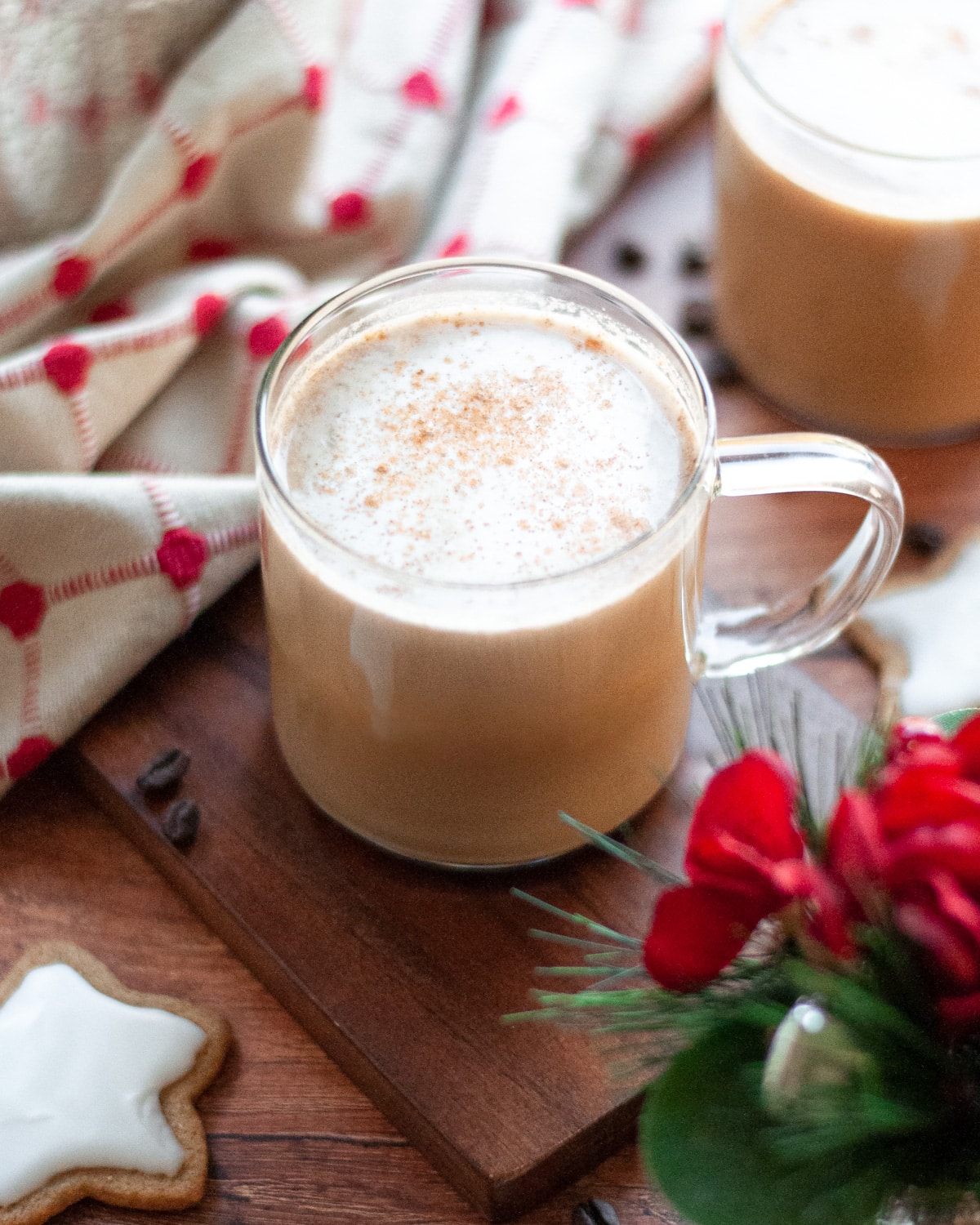 a glass mug with a homemade gingerbread latte sitting on a wooden board. Another mug sits in the background, along with a linen, coffee beans, an iced gingerbread cookie, and a holiday plant.