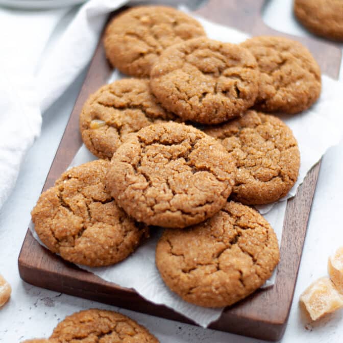 stack of triple ginger molasses cookies on a wooden board with parchment paper. a plate of additional cookies sit next to the board as well as pieces of crystallized ginger and more cookies.