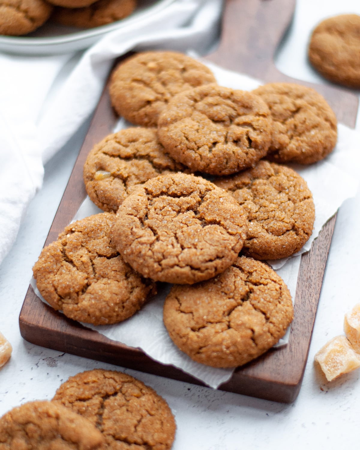 stack of triple ginger molasses cookies on a wooden board with parchment paper. a plate of additional cookies sit next to the board as well as pieces of crystallized ginger and more cookies.