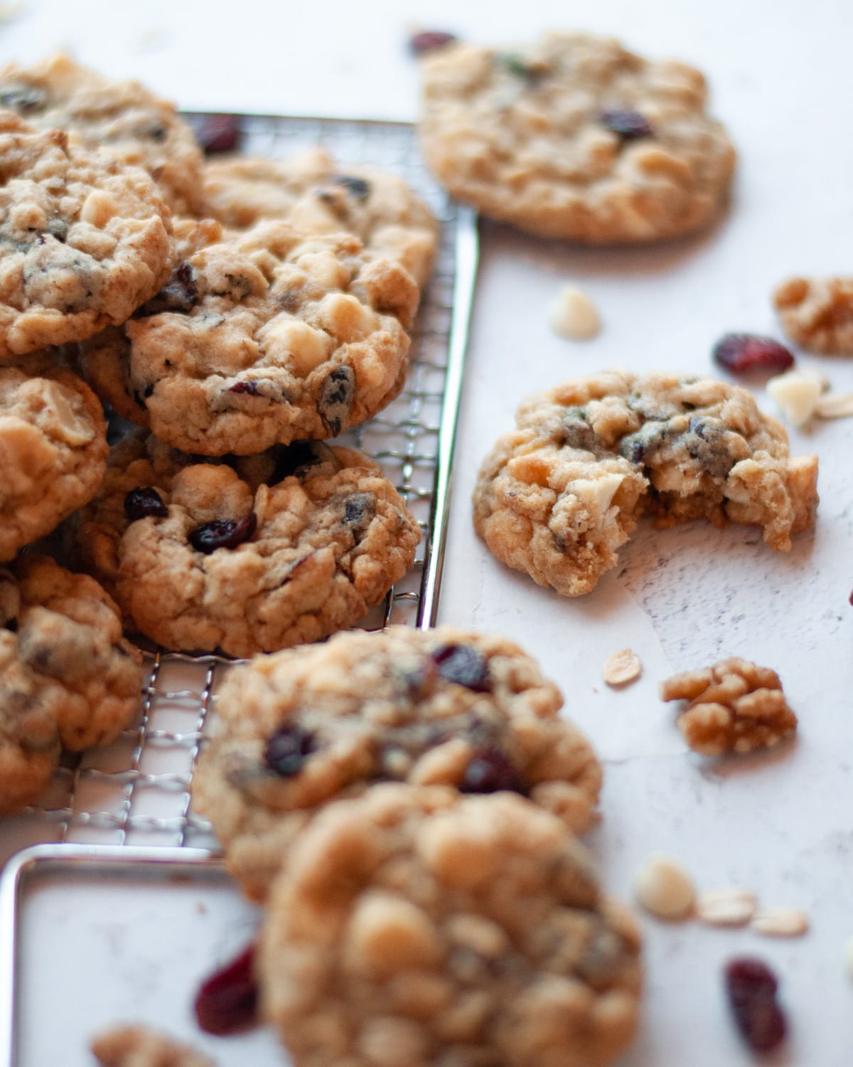 stack of white chocolate cranberry oatmeal cookies on a metal grater. the featured cookie has a bite taken out of it, and walnuts, oatmeal, white chocolate chips, and craisins scattered around.
