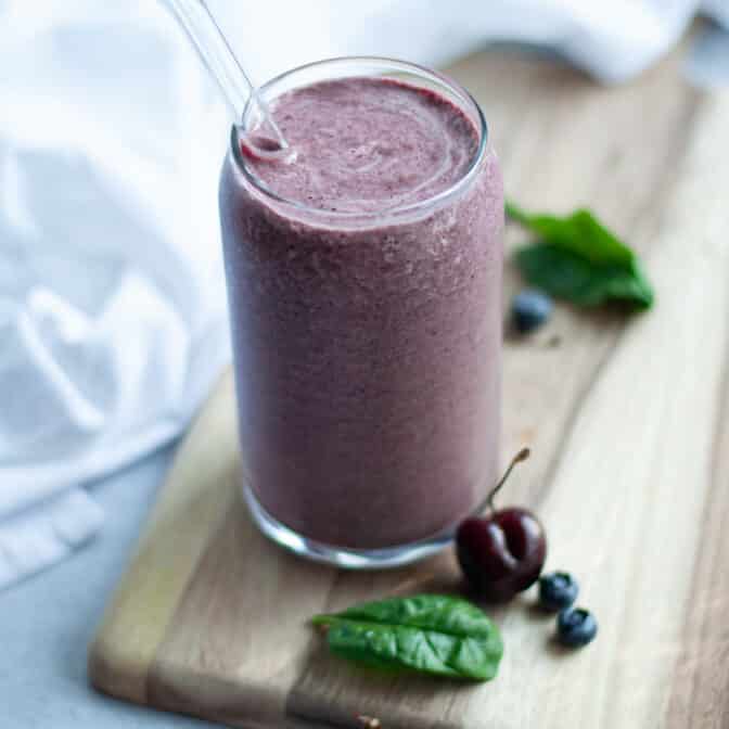 close up of a glass filled to the brim with this blueberry cherry smoothie with spinach. A glass straw is in the glass and ingredients are scattered around.